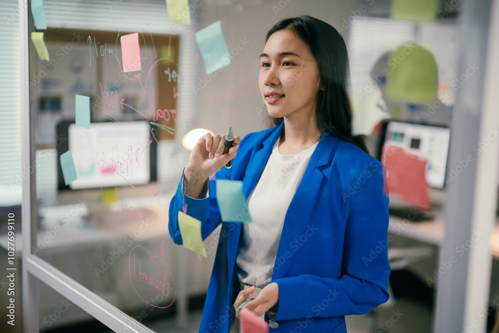 Poster young manager is using colorful sticky notes and a marker to brainstorm ideas on a transparent glass