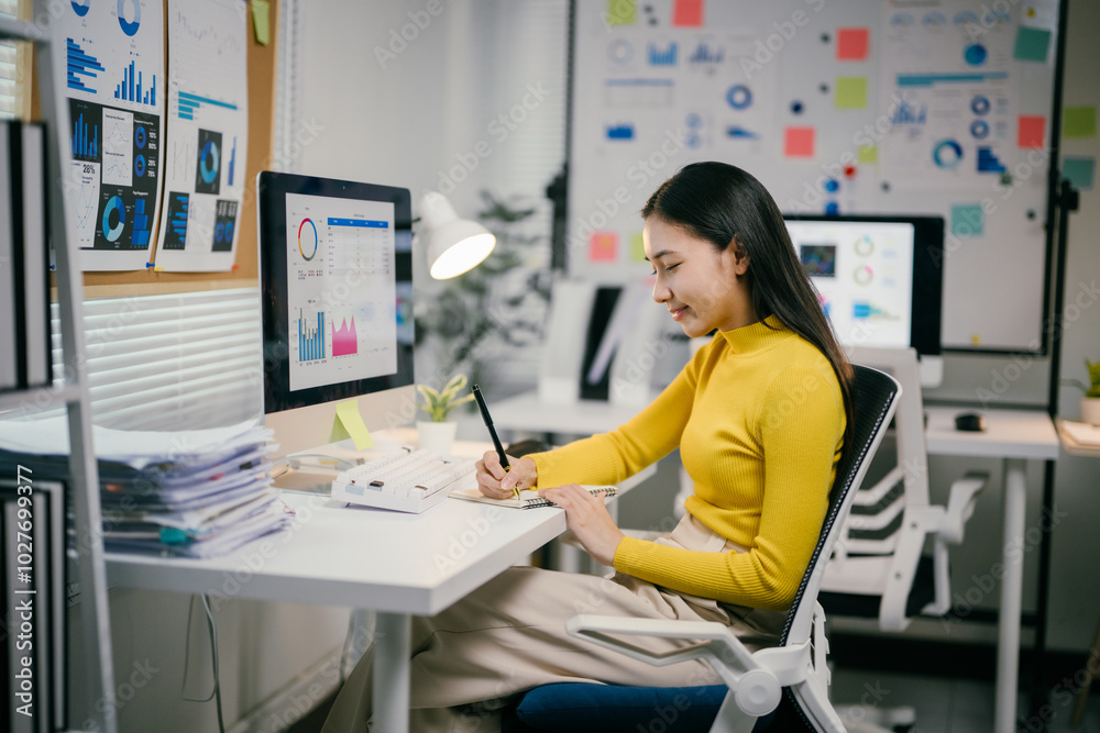 Poster businesswoman is analyzing data charts on her computer and writing notes in her notebook. she is wor