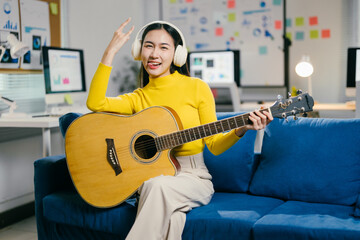 Young asian woman enjoys playing acoustic guitar while taking a break in her home office, wearing...