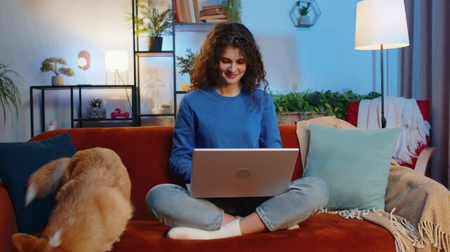 Happy woman with curly hair opening laptop and working remotely sitting beside corgi dog on sofa in living room. Beautiful female girl freelancer in casual clothes smiling and using netbook at home.