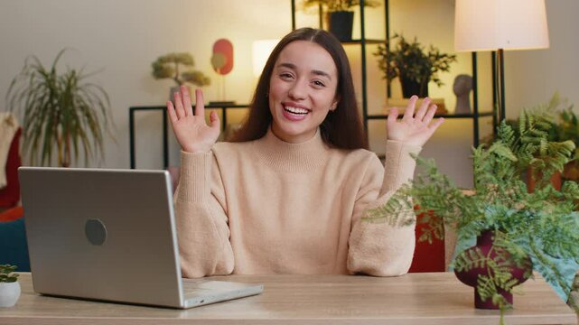 Hello. Pretty young woman smiling friendly at camera, waving hands gesturing hello hi, greeting or goodbye welcoming with hospitable expression at home office. Caucasian girl sitting at table in room