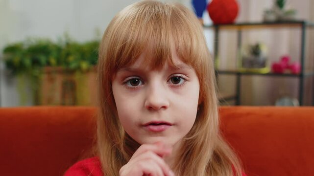 Shh be quiet please. Preteen child girl presses index finger to lips makes silence hush gesture sign do not tell gossip secret. Teenager female little kid at home apartment living room sits on couch