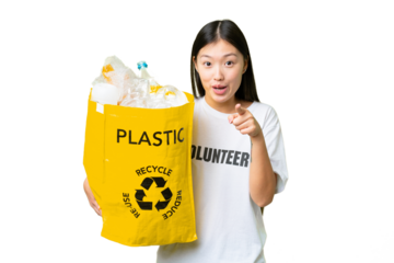Young Asian woman holding a bag full of plastic bottles to recycle over isolated chroma key background surprised and pointing front