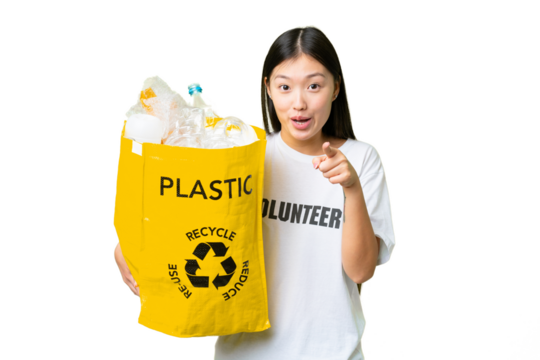 Young Asian woman holding a bag full of plastic bottles to recycle over isolated chroma key background surprised and pointing front - Powered by Adobe