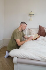 Fototapeta premium A man in a green shirt is kneeling on the floor next to a bed, engrossed in a book. He is surrounded by the warmth and comfort of his home, enjoying a quiet moment of reading.