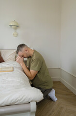 A man kneels in prayer beside a bed, head bowed in devotion. The simple setting emphasizes the sincerity and peacefulness of his moment of reflection.