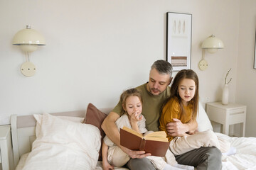 A father sits on a bed, reading a book to his two daughters. The girls are attentively listening, creating a heartwarming moment of togetherness and shared love for literature