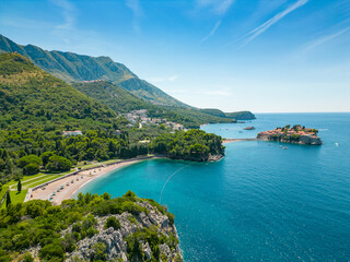 Aerial drone view of Sveti Stefan Island- Luxury travel destination in Montenegro. Waves splashing the rocks. At right is Sveti Stefan Beach. Panoramic view. Emerald water.