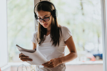 A young woman wearing headphones and glasses reads papers by a window. She is focused and appears...