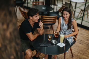 A young couple is seen laughing and chatting at an outdoor cafe table, enjoying cold drinks in a relaxed and friendly atmosphere. Their interaction conveys joy and connection.