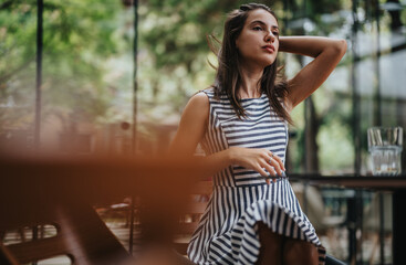 A young woman sits in a modern cafe, wearing a chic striped dress. She looks relaxed and contemplative amidst a serene setting with natural light.