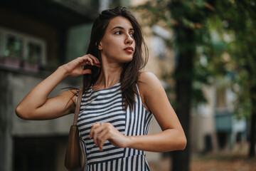 A young woman wearing a striped dress stands outdoors, exuding a thoughtful and reflective demeanor. The blurred background adds depth and urban context to the portrait.