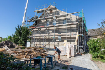 Baugerüst vor einem Haus, bei dem das Dach erneuert wird. Ein Dachdecker sägt eine Latte zurecht.