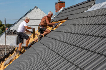 Ein Dach wird neu gedeckt. Beton-Dachsteine in der Farbe Anthrazit. Zwei Dachdecker im Seniorenalter