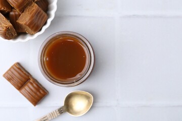 Tasty caramel sauce in jar, spoon and candies on white tiled table, flat lay. Space for text