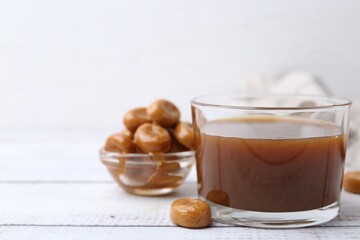 Tasty caramel sauce in bowl and candies on white wooden table, closeup