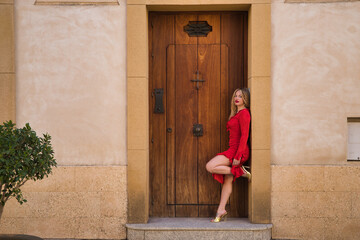 Young, beautiful, blonde woman in red dress and golden heels, posing looking at camera, leaning against a beautiful door frame. Concept beauty, fashion, femininity, trendy.