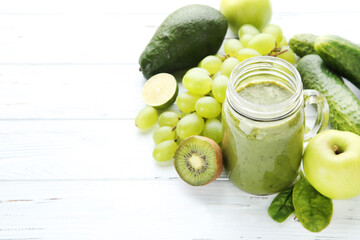 Green smoothie in glass jar with fruits and vegetables on white wooden background