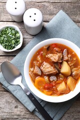 Delicious stew with vegetables in bowl, parsley and spoon on wooden table, top view