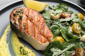 Tasty grilled salmon steak with lemon, sauce and salad on plate, closeup