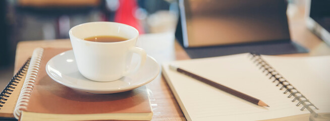 White coffee cup, planner book, stack of notebooks on a wooden desk. Pencil lies on the top notebook, and a tablet and laptop in the background-banner image.