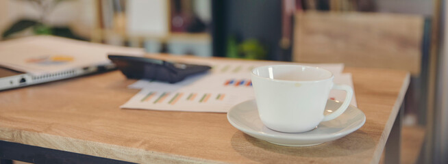 White coffee cup on business wooden desk. Laptop, calculator, report, and papers are blurred in the background at office-banner image.