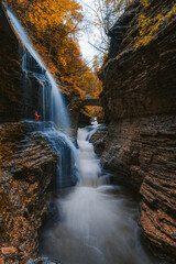 Female Hiker in Orange Coat Under Waterfall. Stone Bridge Over River Watkins Glen State Park. Autumn Season Incredible Canyon Gorge Fantasy Location