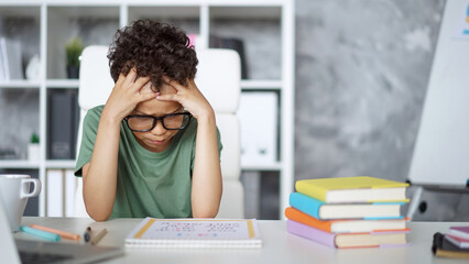  Portrait of a stressed African American schoolboy sitting at the desk holding his head with hands,...
