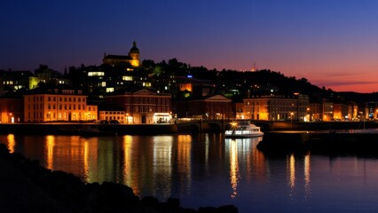 Obraz premium Coastal city at dusk with illuminated buildings reflecting on the water during sunset