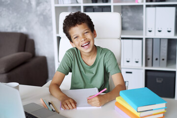 Portrait of adorable happy smiling beautiful kid student joyful schoolchild sitting at the office desk and working on his home assignment