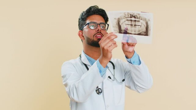 Indian young doctor orthodontist man examines a panoramic x-ray picture of the jaw teeth. 3D model of the patient's mouth, MRI scan. Dentistry, oral care. Arabian stomatology guy on beige background