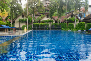 Tropical resort swimming pool with clear blue water, surrounded by lush greenery, palm trees, sun loungers and wooden bungalows, Phi Phi island, Thailand. Luxury travel and tourism