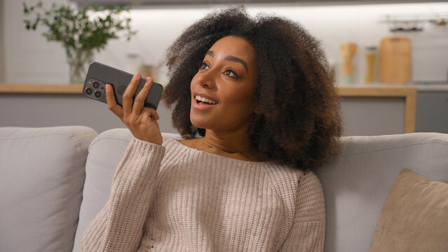 Happy African American woman on couch ethnic smiling female girl mobile phone send audio message using cell speakerphone convenient usage smartphone record voice mail recording chat voicemail at home