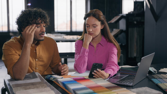 Two designers male female colleagues in modern design studio looking at fabric samples discussing color textile for interior angry Arabian Indian man talk with Caucasian woman problem misunderstanding