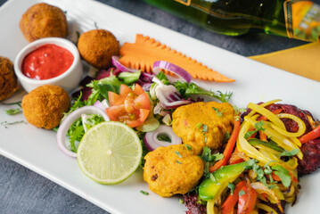 Colorful platter featuring crispy vegetarian falafel balls accompanied by fresh salad greens, red onions, and diced tomatoes served with side of tangy tomato sauce sliced lemon & cilantro.