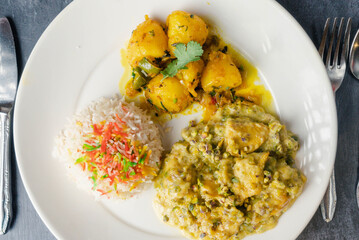 Deshi plate of fluffy colorful rice topped with bits of red and yellow, with golden boiled potatoes seasoned with herbs, greenish chciken curry with vegetables.