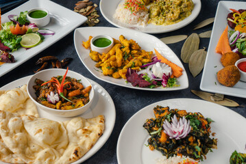 Indian dishes are plated with colorful curries, garnished rice, breads like naan fresh herbs, lime wedges, and onion rings. Side dishes of crispy pakoras, and a fresh salad.
