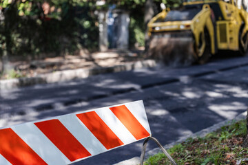 A red and white barricade stands on a newly paved road, with a road roller in the background