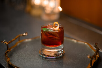 Glass with a brown drink, an ice cube, a piece of zest and a green leaf