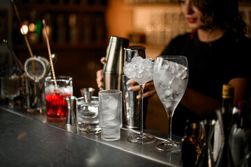 Cute female bartender puts two cocktail shakers on the bar counter, next to which there are various glasses