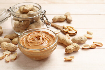 Tasty peanut butter in bowl, groundnuts, jar and spoon on wooden table, closeup