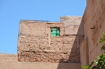 Aged adobe building with a small green window, casting shadows against a bright blue sky in Marrakech
