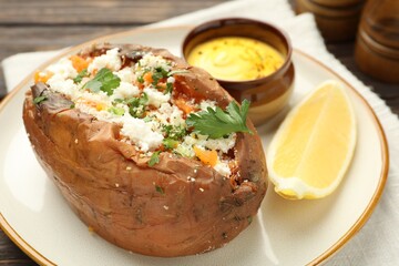 Tasty baked sweet potato with feta cheese, parsley, sauce and lemon slice on table, closeup