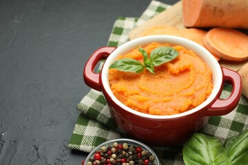 Tasty mashed sweet potato with basil in pot, cut vegetable and spices on dark textured table, closeup. Space for text