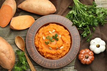Tasty mashed sweet potato with pumpkin seeds in bowl, spoon, fresh vegetables and parsley on wooden table, flat lay