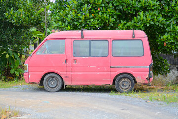 Fototapeta premium A rusty red van is parked on a dirt road, surrounded by lush greenery.