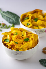 Unripe Plantain chips mixed with herbs in white ceramic bowls on a white background littered with kale, almond nuts parsley and basil