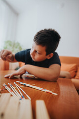 A young boy focused on a game of pick-up sticks, sitting at a wooden table indoors, conveying...
