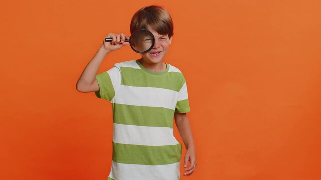 Investigator researcher scientist preteen child boy kid holding magnifying glass near face, looking into camera with big zoomed funny eye, searching, analyzing. Young children on orange background