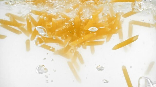 Closeup view of penne pasta thrown into water filmed in slow motion against white background. Macro footage of flour products falling in transparent clear aqua. Concept of cooking food. Eating theme